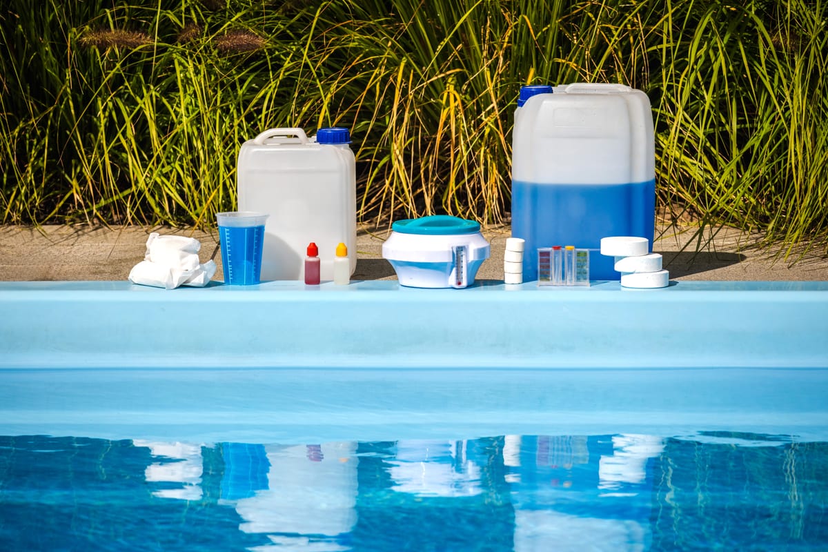 Pool chemical supplies including chlorine tablets, liquid chlorine, acid, and a measuring cup at the edge of a swimming pool